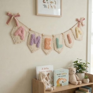Op de afbeelding: Een slinger met de naam "AMELIA" in pastelkleurige letters hangt aan een muur. De slinger is gemaakt van crèmekleurige stof met een kanten rand en is versierd met roze strikken. Een houten plank eronder toont boeken, een knuffelkonijn, een teddybeer en planten.