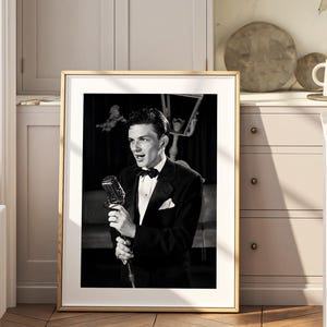 Pode incluir: Uma fotografia emoldurada a preto e branco apresentando uma pessoa de smoking a cantar num microfone vintage. O retrato é exibido numa moldura de madeira clara, capturando um momento de atuação clássica com iluminação de alto contraste.