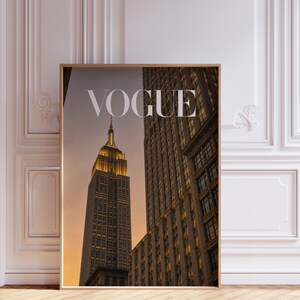 May include: A rectangular wall art print featuring a photograph of the Empire State Building at sunset with the word VOGUE printed in white capital letters at the top. The skyscraper is illuminated with warm golden lights against a dusk sky.