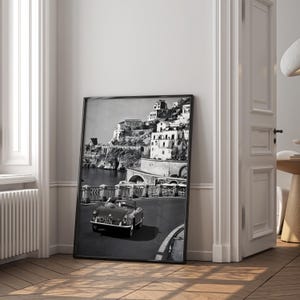 May include: Monochrome framed print depicting a coastal town with buildings on a hillside. A classic convertible car is on a road in the foreground. The print is propped against a white wall in a room with a wooden floor.