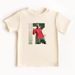 Puede incluir: Camiseta color crema con un gr&aacute;fico de un futbolista con camiseta roja con el n&uacute;mero 7, pantalones cortos verdes y una firma. La palabra "RONALDO" est&aacute; impresa verticalmente en verde y rojo.