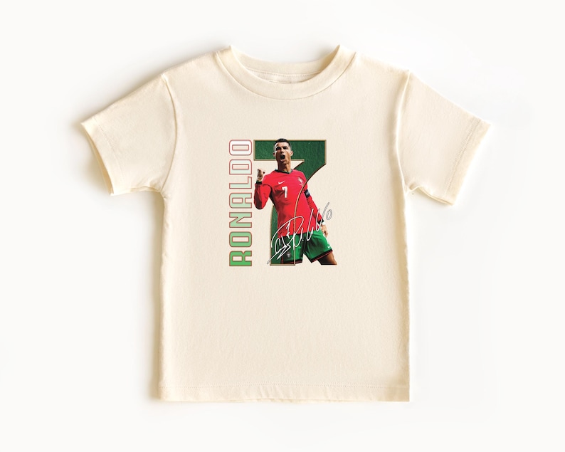 Puede incluir: Camiseta color crema con un gr&aacute;fico de un futbolista con camiseta roja con el n&uacute;mero 7, pantalones cortos verdes y una firma. La palabra "RONALDO" est&aacute; impresa verticalmente en verde y rojo.