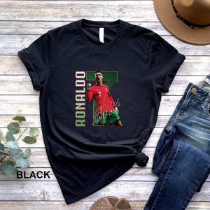 Puede incluir: Camiseta negra con un gr&aacute;fico de un futbolista con camiseta roja, el nombre "Ronaldo" en texto vertical verde y rojo, y una firma. El jugador se muestra celebrando. La palabra "BLACK" est&aacute; impresa en la parte inferior.