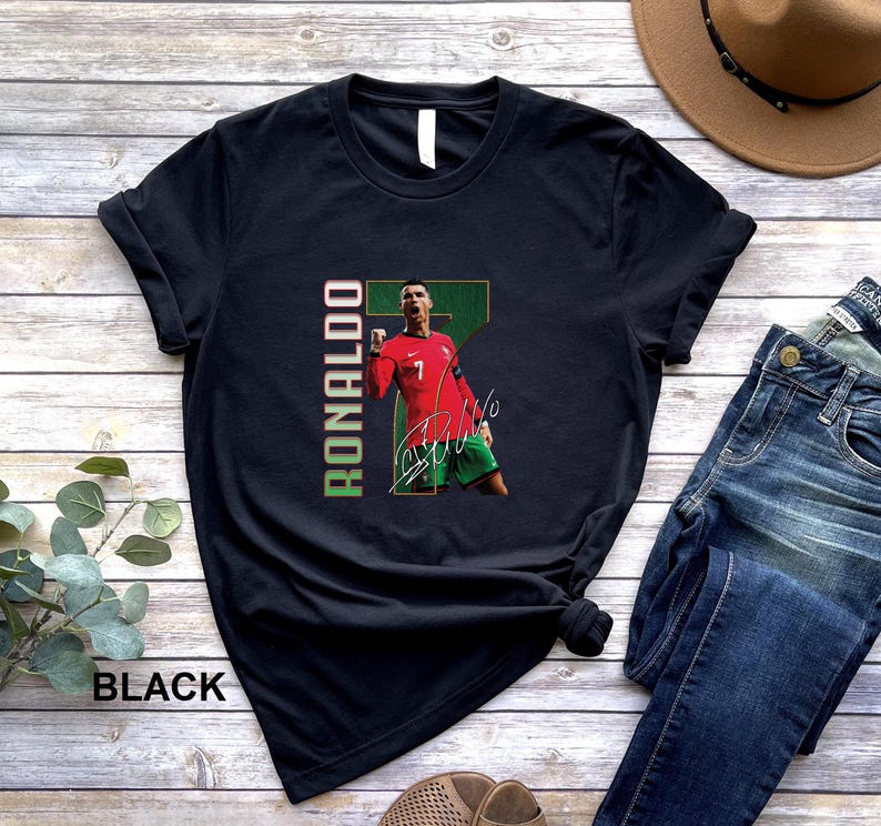 Puede incluir: Camiseta negra con un gr&aacute;fico de un futbolista con camiseta roja, el nombre "Ronaldo" en texto vertical verde y rojo, y una firma. El jugador se muestra celebrando. La palabra "BLACK" est&aacute; impresa en la parte inferior.