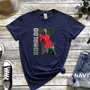 Puede incluir: Camiseta azul marino con un gr&aacute;fico de un jugador de f&uacute;tbol con una camiseta roja con el n&uacute;mero 7 y el nombre "RONALDO" en verde y rojo. La camiseta tambi&eacute;n incluye la firma del jugador. La palabra "NAVY" est&aacute; impresa en los vaqueros.