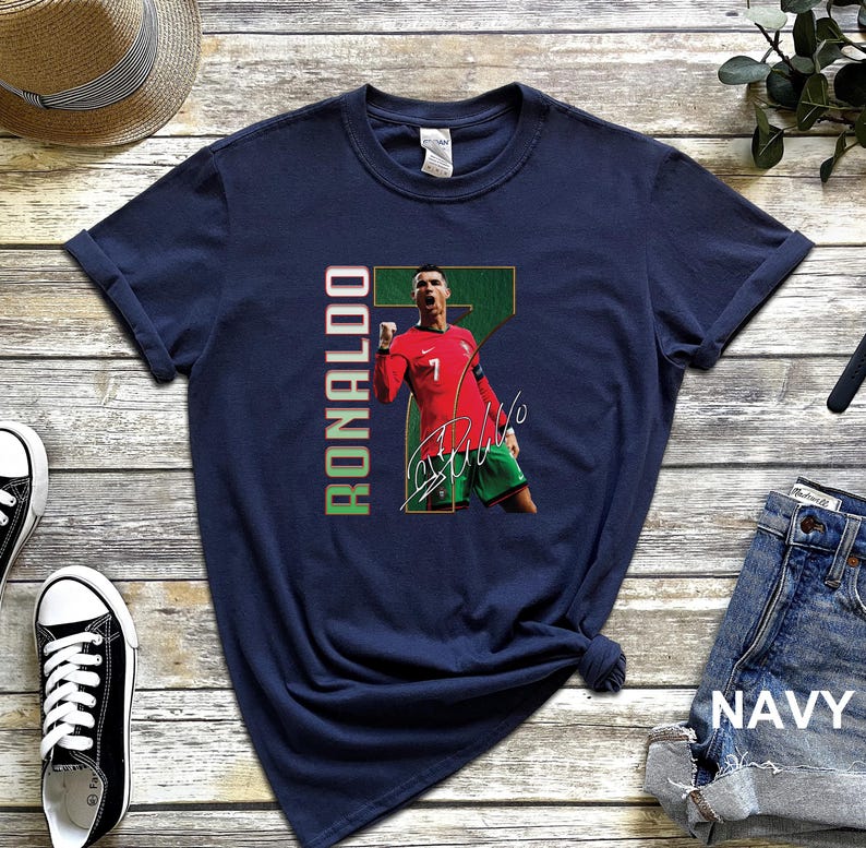 Puede incluir: Camiseta azul marino con un gr&aacute;fico de un jugador de f&uacute;tbol con una camiseta roja con el n&uacute;mero 7 y el nombre "RONALDO" en verde y rojo. La camiseta tambi&eacute;n incluye la firma del jugador. La palabra "NAVY" est&aacute; impresa en los vaqueros.