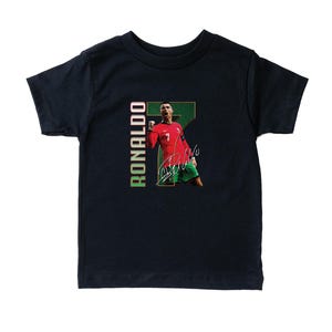 Puede incluir: Camiseta azul marino para ni&ntilde;os peque&ntilde;os con un gr&aacute;fico de un jugador de f&uacute;tbol con una camiseta roja con el n&uacute;mero 7. La camiseta tiene el nombre "RONALDO" impreso verticalmente en verde y blanco, junto con la firma del jugador.