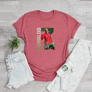 Puede incluir: Una camiseta roja con un gr&aacute;fico de un jugador de f&uacute;tbol con uniforme rojo, el n&uacute;mero 7, el nombre "RONALDO" en verde y una firma. La camiseta se muestra sobre una superficie de color claro.