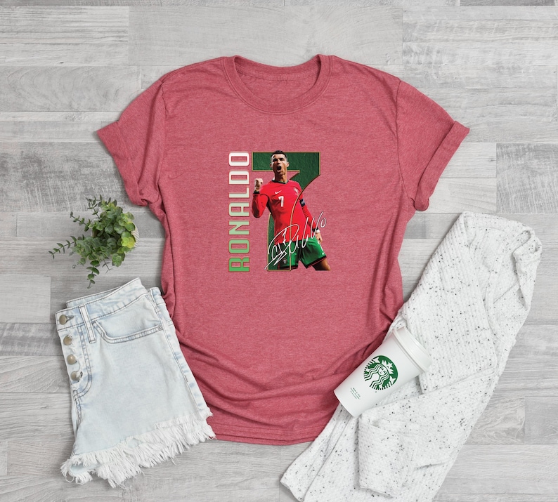 Puede incluir: Una camiseta roja con un gr&aacute;fico de un jugador de f&uacute;tbol con uniforme rojo, el n&uacute;mero 7, el nombre "RONALDO" en verde y una firma. La camiseta se muestra sobre una superficie de color claro.