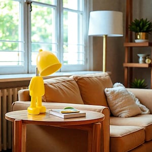 May include: A quirky yellow desk lamp, shaped like a person with a rounded head and legs, stands on a wooden table. A book rests beside it. The background features a window, a sofa, and a floor lamp, creating a cozy interior scene.