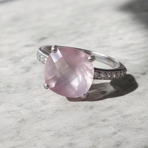 May include: A silver ring featuring a large, faceted, light pink gemstone. The band is also adorned with smaller, clear gemstones. The ring is set against a light gray, textured surface.