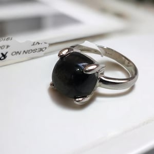 Pode incluir: Um anel de prata com uma pedra preciosa preta grande e arredondada. A pedra preciosa é mantida no lugar por quatro garras de prata. O anel está sobre uma superfície branca. A etiqueta ao lado do anel tem o texto "DESIGN NO R-72".