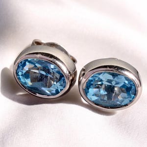 May include: A pair of silver-toned stud earrings, each featuring a large, oval-cut, light blue gemstone. The gemstones are set in a bezel setting, and the earrings are displayed on a white, textured surface.