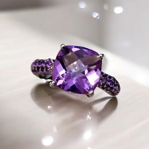 May include: A silver ring with a large, square-cut, purple gemstone as the centerpiece. Smaller, round purple stones adorn the ring's band on either side of the main stone. The ring is set against a white surface.
