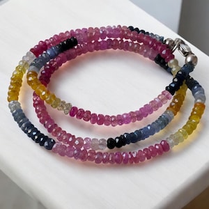 Puede incluir: Un collar de varias hebras compuesto por pequeñas piedras preciosas facetadas en una variedad de colores, incluyendo rosa, amarillo, azul y negro. El collar está asegurado con un cierre plateado y se muestra sobre una superficie blanca.