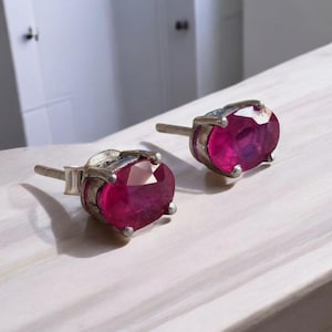 K&ouml;nnte beinhalten: Ein Paar silberne Ohrstecker, jeweils mit einem gro&szlig;en, oval geschliffenen Rubin. Die Rubine sind tiefrot und in einer silbernen Fassung gefasst. Die Ohrringe werden auf einer wei&szlig;en Oberfl&auml;che pr&auml;sentiert.