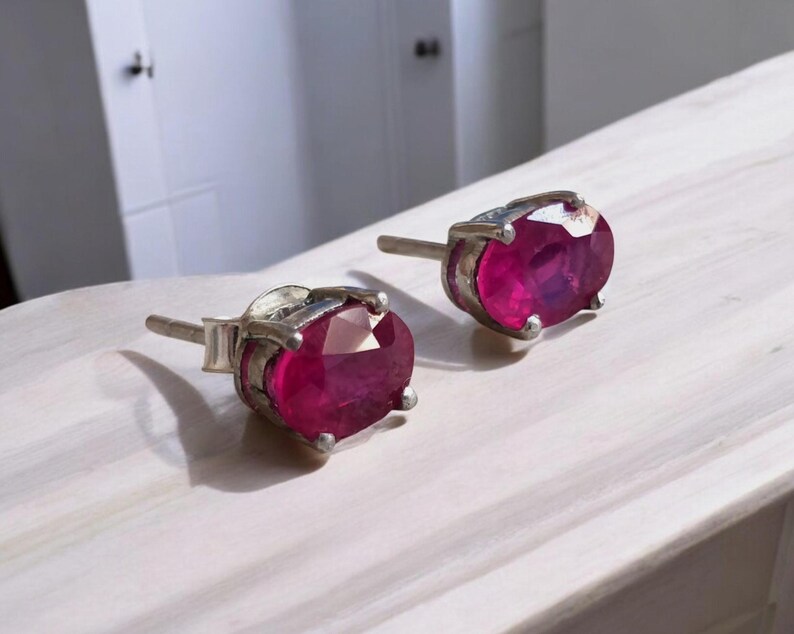K&ouml;nnte beinhalten: Ein Paar silberne Ohrstecker, jeweils mit einem gro&szlig;en, oval geschliffenen Rubin. Die Rubine sind tiefrot und in einer silbernen Fassung gefasst. Die Ohrringe werden auf einer wei&szlig;en Oberfl&auml;che pr&auml;sentiert.