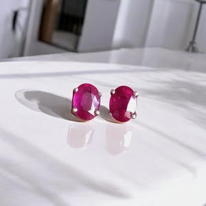 Op de afbeelding: Een paar ovale robijn oorknopjes. De robijnen hebben een diepe, levendige rode kleur en zijn gezet in een zilverkleurig metaal. De oorbellen worden gepresenteerd op een wit oppervlak.
