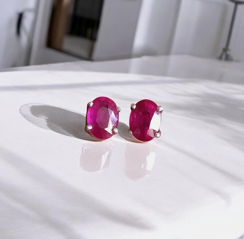 K&ouml;nnte beinhalten: Ein Paar ovale Rubin-Ohrstecker. Die Rubine haben eine tiefe, lebendige rote Farbe und sind in einem silberfarbenen Metall gefasst. Die Ohrringe werden auf einer wei&szlig;en Oberfl&auml;che pr&auml;sentiert.