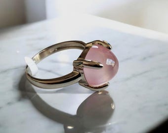 Puede incluir: Un anillo de plata con una gema grande, redonda y rosa claro. La gema está sujeta por cuatro garras de plata. El anillo está sobre una superficie blanca, con una sombra proyectada.