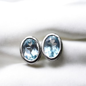 May include: A pair of oval-shaped stud earrings. Each earring features a light blue gemstone set in a silver-colored metal frame. The gemstones have a faceted cut, reflecting light. The earrings are displayed on a white, textured background.