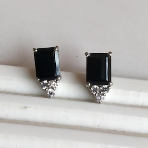 May include: A pair of stud earrings featuring rectangular black gemstones. Each earring has a silver-colored setting with a cluster of small, clear gemstones at the base, creating a sparkling accent. The earrings are displayed on a white surface.