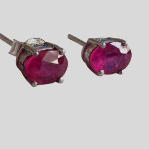 K&ouml;nnte beinhalten: Ein Paar silberne Ohrstecker, jeweils mit einem oval geschliffenen Rubin. Die Rubine haben eine tiefrote Farbe und sind in einer vierzackigen Silberfassung gefasst. Die Ohrringe sind vor einem hellgrauen Hintergrund abgebildet.