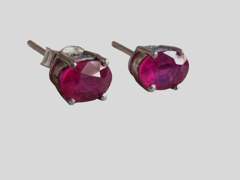 K&ouml;nnte beinhalten: Ein Paar silberne Ohrstecker, jeweils mit einem oval geschliffenen Rubin. Die Rubine haben eine tiefrote Farbe und sind in einer vierzackigen Silberfassung gefasst. Die Ohrringe sind vor einem hellgrauen Hintergrund abgebildet.