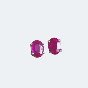 K&ouml;nnte beinhalten: Ein Paar ovale Rubin-Ohrstecker. Die Rubine haben eine leuchtende magenta Farbe und sind in einem silberfarbenen Metall gefasst. Die Ohrringe werden vor einem schlichten wei&szlig;en Hintergrund pr&auml;sentiert.