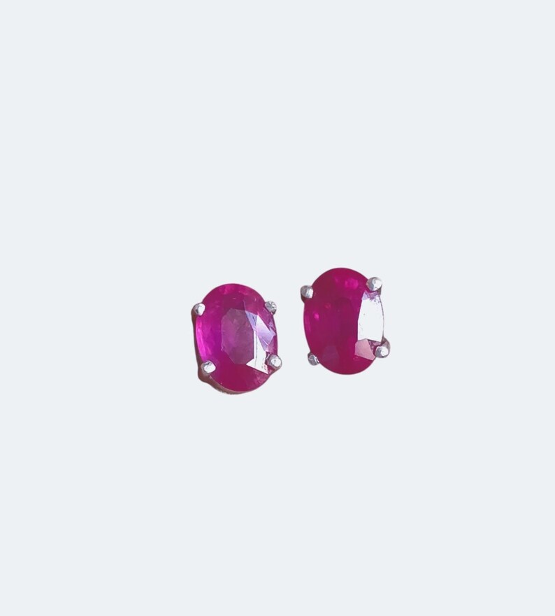 K&ouml;nnte beinhalten: Ein Paar ovale Rubin-Ohrstecker. Die Rubine haben eine leuchtende magenta Farbe und sind in einem silberfarbenen Metall gefasst. Die Ohrringe werden vor einem schlichten wei&szlig;en Hintergrund pr&auml;sentiert.