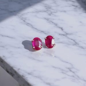 K&ouml;nnte beinhalten: Ein Paar Ohrringe mit oval geschliffenen Rubinen. Jeder Ohrring hat einen leuchtend tiefroten Edelstein, der in einer Silber- oder Wei&szlig;goldeinfassung gefasst ist. Die Ohrringe werden auf einer wei&szlig;en Marmoroberfl&auml;che pr&auml;sentiert.