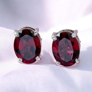May include: A pair of oval-shaped stud earrings. Each earring features a large, deep red gemstone set in a silver-colored metal. The gemstones have a polished, reflective surface. The earrings are displayed on a soft, light pink background.