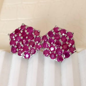 May include: A pair of hexagonal stud earrings. Each earring is composed of numerous small, round, deep pink gemstones set in a silver-colored metal. The gemstones are closely packed together, creating a textured surface. The earrings are displayed against a light background.