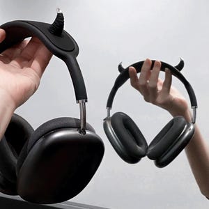 May include: Black over-ear headphones with a devil horn accessory. The headphones have a sleek, modern design with a dark grey mesh ear cup and a silver metal band. The devil horn accessory is black and adds a unique touch.