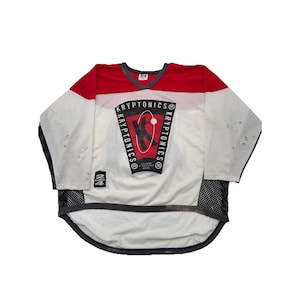 May include: White and red vintage hockey jersey with the word "KRYPTONICS" in a circular design on the front. The jersey has a V-neck and black mesh detailing on the sleeves and bottom hem. The jersey has a few stains.
