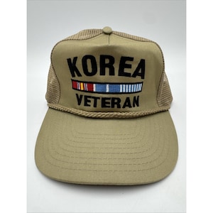 May include: A tan baseball cap with a mesh back and a braided cord trim across the front. The front panel features the black embroidered text KOREA VETERAN with a colorful ribbon bar graphic between the two words, all centered on the cap.