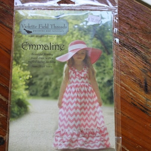 May include: A package for a sewing pattern titled "Emmaline" by Violette Field Threads. The pattern features a girl wearing a pink and white chevron maxi dress and a matching sun hat. The text on the package says "Beautiful flowing maxi dress with a ruffled halter neckline. Sizes 2T-10 Years."