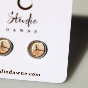May include: A pair of silver stud earrings with a white background and a clock face design. The earrings are on a white card with the text "Studio Dawne" and "studioodawne.com".
