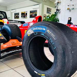 Può includere: Uno pneumatico da corsa liscio nero Michelin con il logo blu e giallo, la mascotte Bibendum e la scritta TRJ-1 stampata sul fianco. Lo pneumatico è posizionato davanti a un'auto da corsa di Formula 1 rossa e arancione in un garage.