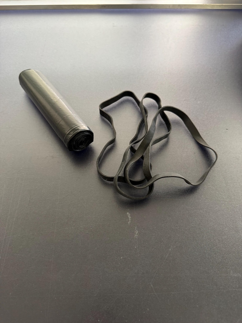 May include: A black roll of plastic trash bags and several black rubber bands are on a dark surface. The bags are cylindrical and tightly rolled. The rubber bands are looped and scattered.