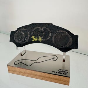 Peut inclure: Une pièce décorative noire avec trois motifs circulaires et le numéro "304" en jaune. Elle est montée sur une base en bois avec une plaque argentée représentant un tracé de circuit et le texte "ASSEN TT CIRCUIT".