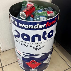 May include: A white and blue metal barrel filled with plastic water bottles and a red soda can. The barrel has the text "It's wonderful! Panta Racing Fuel" printed on it. A red and white hazard label is visible.
