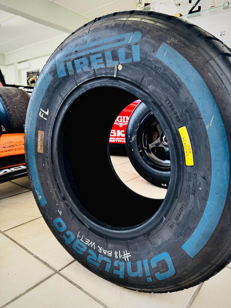 May include: A black Pirelli Cinturato wet racing tire featuring blue lettering on the sidewall. The tire is marked with FL, #18 BAR WET4, and a yellow Driver Academy sticker. The text 305/680-18 is visible on the side of the rubber tire casing.