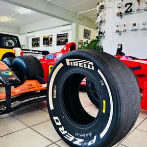 May include: A black Pirelli P Zero Formula 1 racing tire with white lettering on the sidewall. The tire features a yellow sticker, a barcode, and the handwritten text #18 BAK S1. It is positioned in front of a red and orange race car.