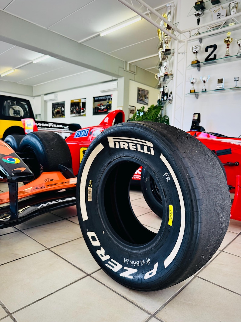 May include: A black Pirelli P Zero Formula 1 racing tire with white lettering on the sidewall. The tire features a yellow sticker, a barcode, and the handwritten text #18 BAK S1. It is positioned in front of a red and orange race car.