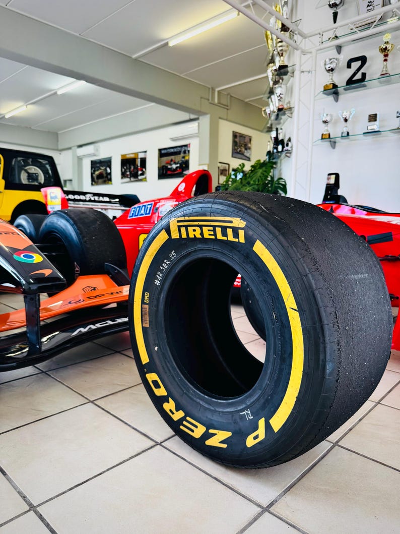 May include: A black Pirelli P Zero Formula 1 racing tire with yellow sidewall lettering, featuring the brand name and model text. The tire is positioned in front of a red and orange race car chassis with visible logos and a smooth, worn tread surface.