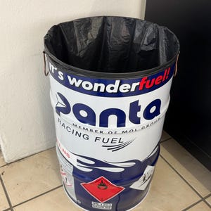 May include: A cylindrical metal trash can with a black plastic liner. The can is white with blue and red graphics, including the word "panta" and "RACING FUEL." A red diamond hazard symbol is visible.