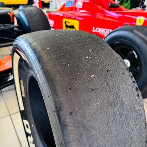 May include: A black racing slick tire with visible wear and debris on the tread surface. The tire features white lettering on the sidewall that reads ZERO. It is positioned in front of a bright red Formula One race car with yellow logos.