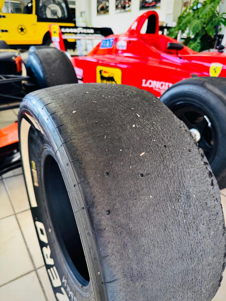 May include: A black racing slick tire with visible wear and debris on the tread surface. The tire features white lettering on the sidewall that reads ZERO. It is positioned in front of a bright red Formula One race car with yellow logos.