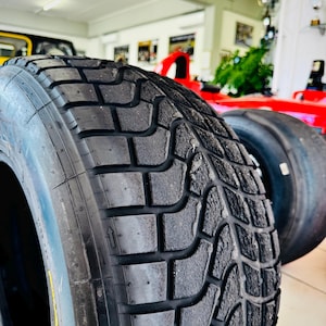 May include: A black rubber tire featuring a deep, intricate tread pattern with rectangular blocks and grooves. The tire surface shows signs of wear and texture. A small directional arrow is embossed on the sidewall, indicating rotation.